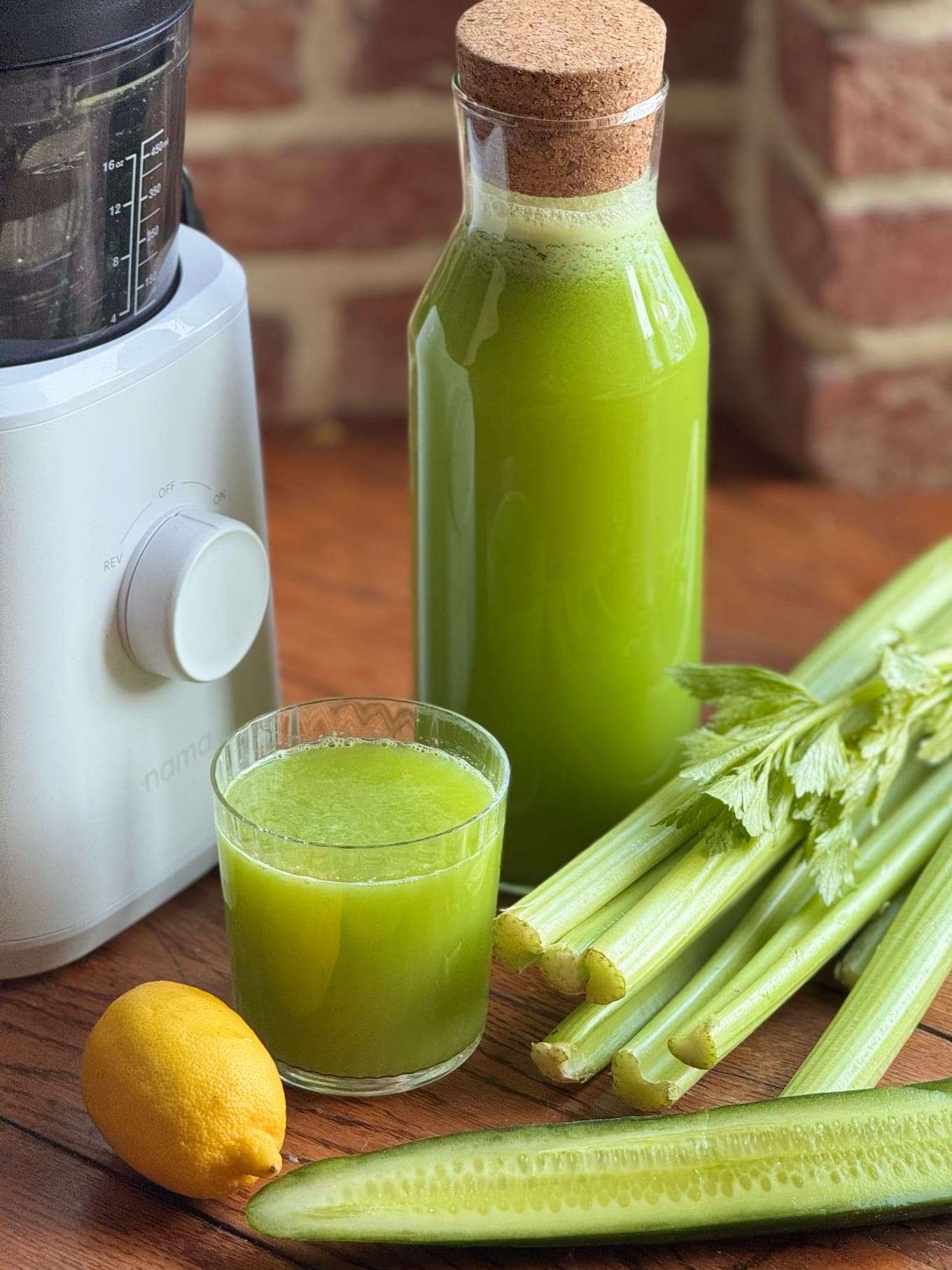 Fresh green juice in a glass and bottle beside a Nama cold press juicer, with celery, cucumber and lemon
